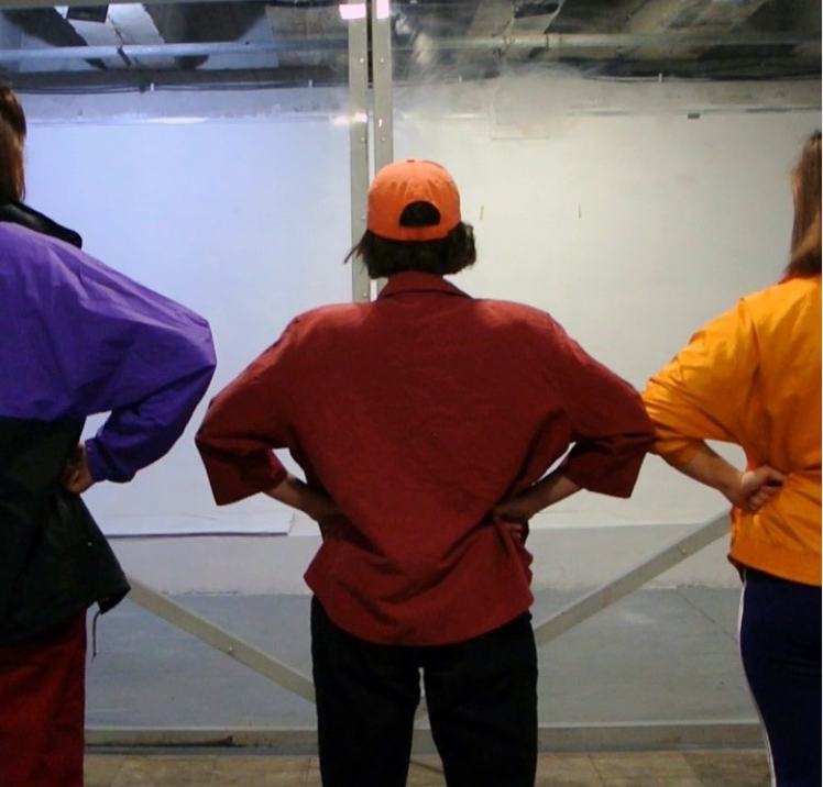three people standing with their back to the camera and hands on their hops in an art studio space. Each wearing a different coloured jumper, purple, red and yellow.